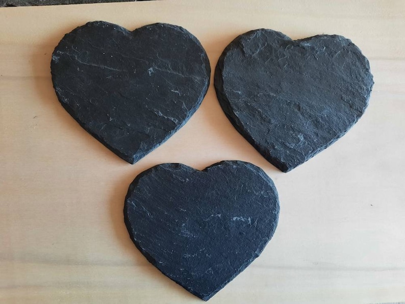 May include: Three black heart-shaped slate coasters. The coasters are made of natural slate and have a rustic, uneven texture.