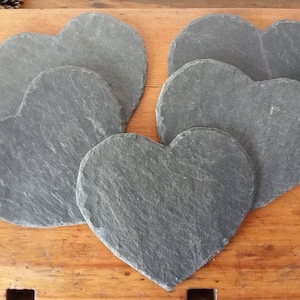 May include: Six gray heart-shaped slate coasters. The coasters are arranged in a stack, with the largest coaster on the bottom and the smallest coaster on top.
