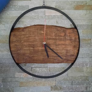 Könnte beinhalten: Ein rustikales Holzuhrblatt mit schwarzen Zeigern, eingefasst in einen schwarzen Metallkreisrahmen. Die Uhr hängt an einer hellgrauen Steinwand.