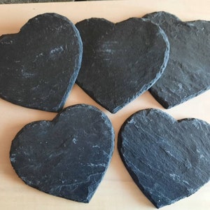 May include: Five black heart-shaped slate coasters. The coasters are made of natural slate and have a rustic, uneven texture.