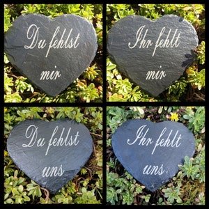 Può includere: Quattro pietre di ardesia a forma di cuore con scritte bianche. Due pietre recitano "Du fehlst mir" e "Ihr fehlt mir", e le altre due recitano "Du fehlst uns" e "Ihr fehlt uns". Le pietre sono su un fogliame verde.