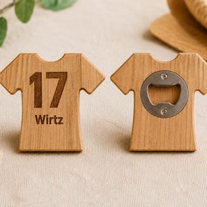 Könnte beinhalten: Zwei hellbraune Flaschenöffner aus Holz in T-Shirt-Form. Der linke Öffner zeigt die eingravierte Zahl 17 und den Namen Wirtz in Dunkelbraun. Der rechte Öffner verfügt über einen runden, silberfarbenen Metall-Flaschenöffner in der Mitte.