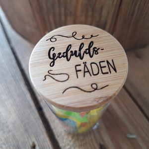 Puede incluir: Un tarro de cristal transparente con tapa de madera grabada con las palabras "Gedulds-Fäden". El tarro contiene notas de papel de colores. El conjunto está sobre un fondo de madera.