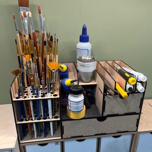 May include: A wooden organizer with multiple compartments for storing art supplies. The organizer has a drawer and a section for holding paintbrushes. There are several paintbrushes, markers, and bottles of glue in the organizer.