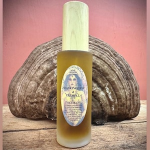 May include: A glass facial oil bottle with a wooden cap, labeled "Frankincense & Tremella Facial Oil." The label features a woman's portrait. The bottle is set against a textured, brown background.