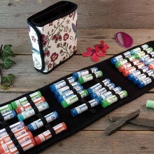 Può includere: Un astuccio nero arrotolabile con più flaconi di vari colori, tra cui blu, verde e arancione. È presente anche un contenitore con stampa floreale. I flaconi sono organizzati in file, suggerendo un kit di rimedi omeopatici.