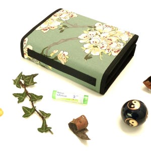 Op de afbeelding: Een rechthoekige, opgevouwen stoffen etui met een bloemenpatroon in groen, wit en bruin. De etui is omgeven door een zwarte rand. Andere items zijn een takje klimop, een klein flesje, een kleine tube, een yin-yang bal en een opgerold bruin object.