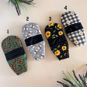 May include: Four different oven mitts with various patterns. The mitts are gray with bees, black with sunflowers, green with butterflies, and black and white gingham with daisies.