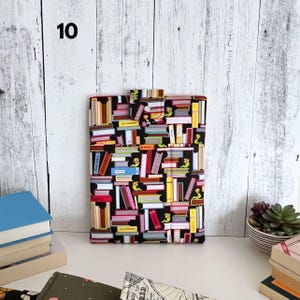 May include: A rectangular fabric sleeve featuring a black background with a colorful pattern of stacked books and small green bookworms. The sleeve includes a small flap closure at the top center and displays the word Dictionary on two spines.