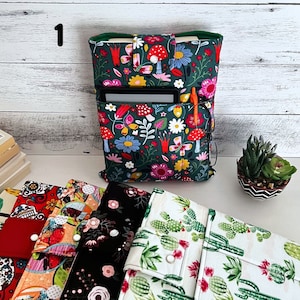 May include: A dark green fabric book sleeve featuring a colorful pattern of red mushrooms, pink and yellow flowers, and butterflies. The sleeve includes a front pocket containing a black e-reader, an orange pen, and black-rimmed glasses.