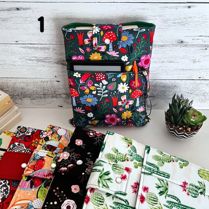 May include: A dark green fabric book sleeve featuring a colorful pattern of red mushrooms, pink and yellow flowers, and butterflies. The sleeve includes a front pocket containing a black e-reader, an orange pen, and black-rimmed glasses.