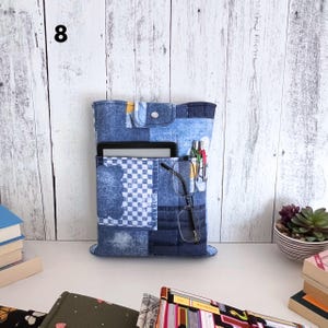 May include: A blue denim fabric organizer sleeve featuring a patchwork design with a checkered pattern, a top flap with a silver snap button, and multiple exterior pockets holding a tablet, two pens, and a pair of black-framed eyeglasses.