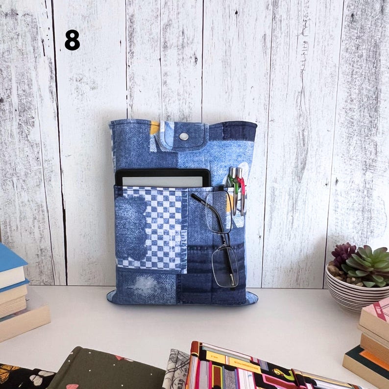 May include: A blue denim fabric organizer sleeve featuring a patchwork design with a checkered pattern, a top flap with a silver snap button, and multiple exterior pockets holding a tablet, two pens, and a pair of black-framed eyeglasses.