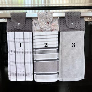 May include: Three kitchen towels hanging on a rack. The towels are gray and white with different patterns. The first towel has a plaid pattern, the second has a striped pattern, and the third has a textured pattern. The towels are numbered 1, 2, and 3.