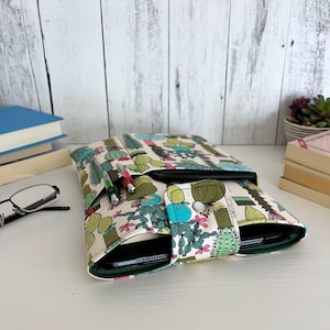 May include: A fabric tablet sleeve featuring a repeating cactus and succulent pattern in shades of green, pink, and yellow on a cream background. The sleeve includes a front pocket with two pens and a snap-button closure flap over the main compartment.