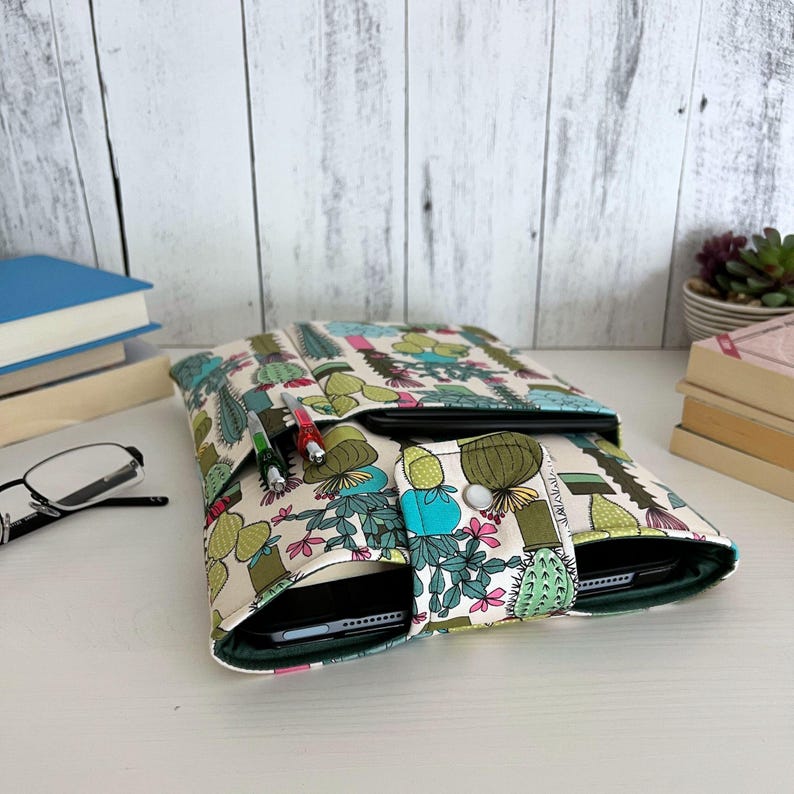 May include: A fabric tablet sleeve featuring a repeating cactus and succulent pattern in shades of green, pink, and yellow on a cream background. The sleeve includes a front pocket with two pens and a snap-button closure flap over the main compartment.