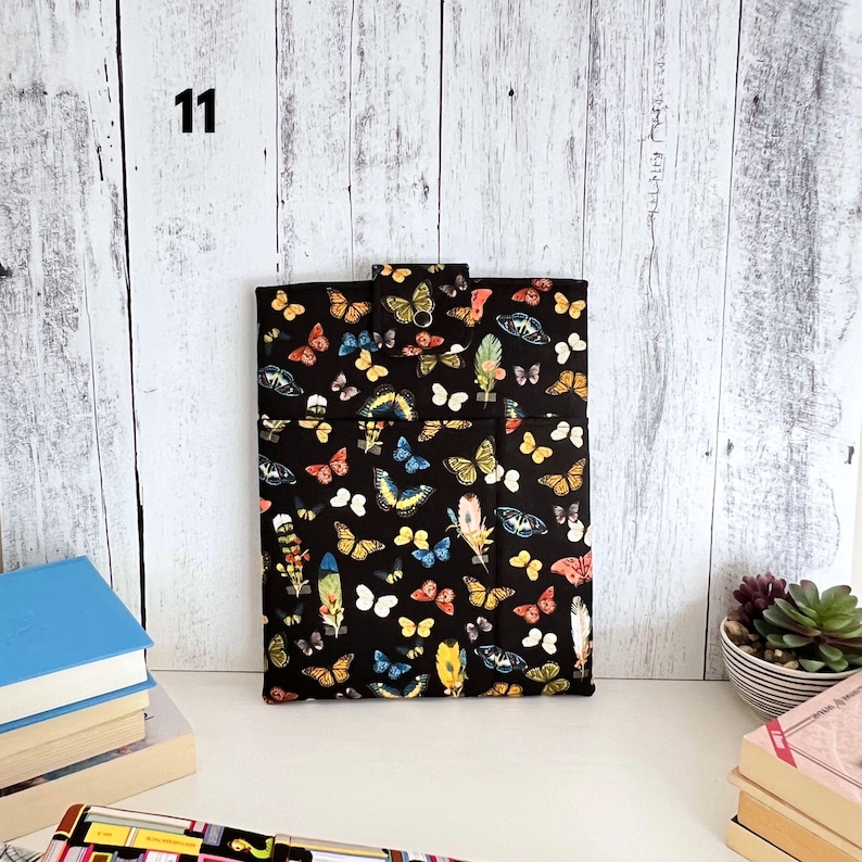 May include: A rectangular fabric sleeve featuring a black background with a repeating pattern of colorful butterflies and feathers. The sleeve includes a top flap with a snap closure and a front pocket, showcasing various species of butterflies.