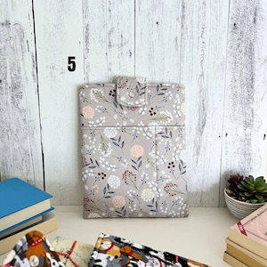 May include: A rectangular fabric sleeve featuring a light gray background with a repeating pattern of small white and pink flowers, dark leaves, and thin stems. The sleeve includes a top flap secured with a silver metal snap button closure.