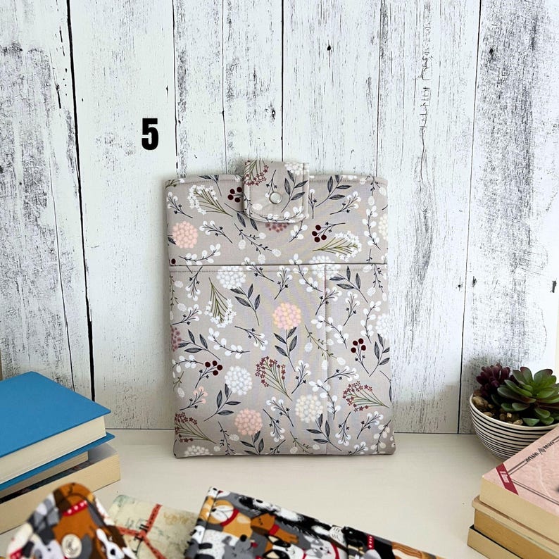May include: A rectangular fabric sleeve featuring a light gray background with a repeating pattern of small white and pink flowers, dark leaves, and thin stems. The sleeve includes a top flap secured with a silver metal snap button closure.