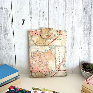 May include: A rectangular fabric sleeve featuring a vintage-style world map print in shades of beige, pink, and cream. The sleeve includes a front pocket and a top flap secured with a single silver-toned metal snap button closure.