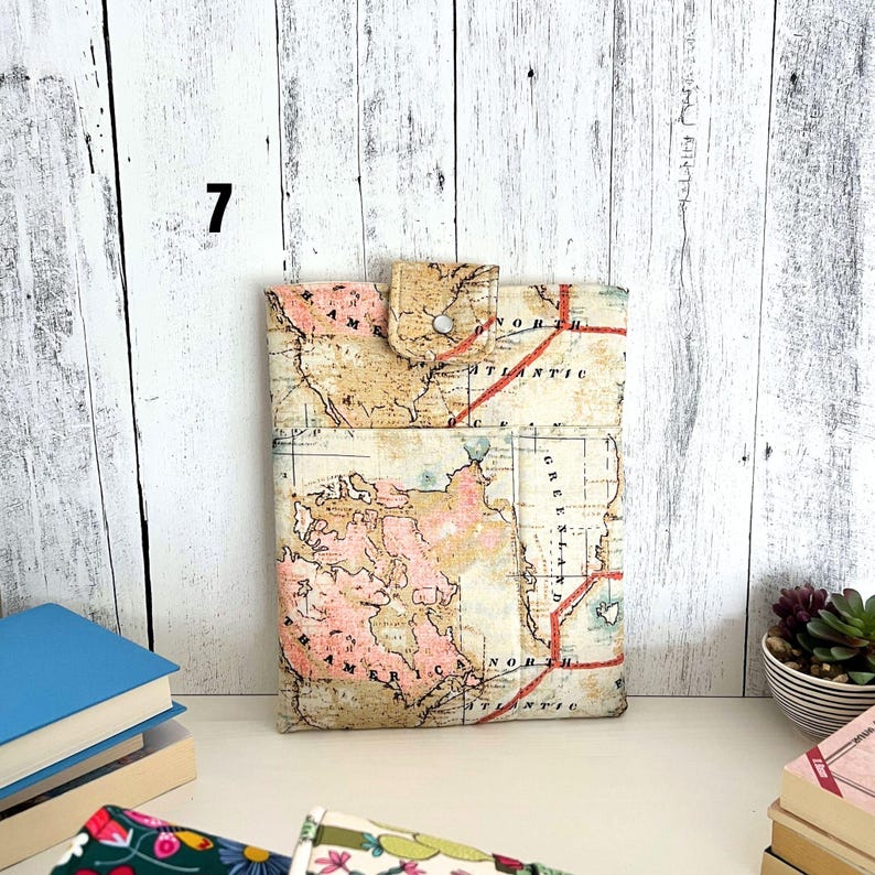 May include: A rectangular fabric sleeve featuring a vintage-style world map print in shades of beige, pink, and cream. The sleeve includes a front pocket and a top flap secured with a single silver-toned metal snap button closure.