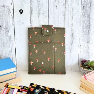May include: A rectangular olive green fabric sleeve featuring a repeating pattern of small red and white mushrooms. The sleeve includes a front pocket and a top flap secured with a white snap button. The number 9 is visible in the upper left corner.