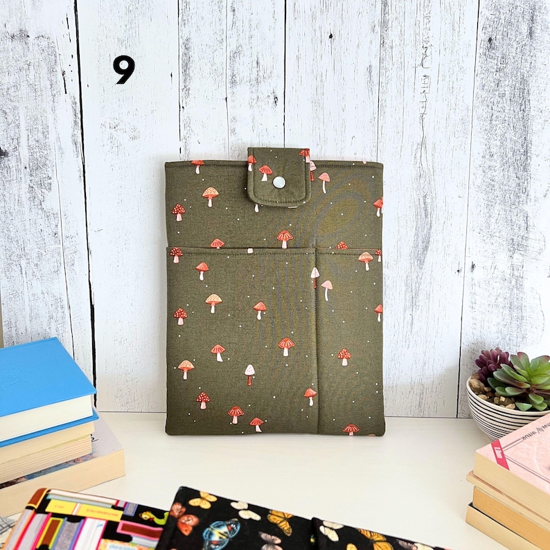 May include: A rectangular olive green fabric sleeve featuring a repeating pattern of small red and white mushrooms. The sleeve includes a front pocket and a top flap secured with a white snap button. The number 9 is visible in the upper left corner.