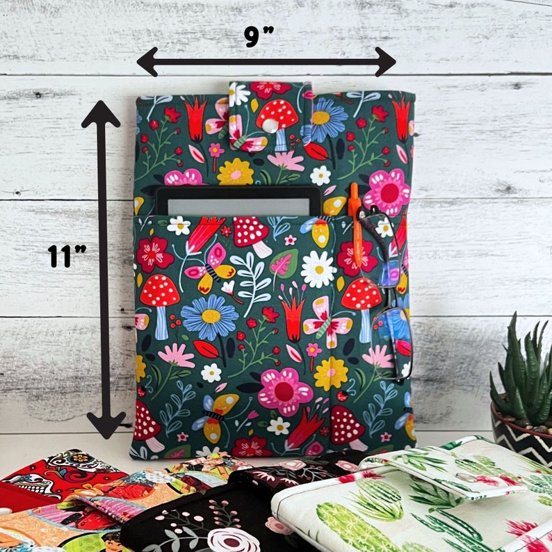 May include: A dark green fabric sleeve featuring a colorful pattern of red mushrooms, yellow and pink flowers, and butterflies. The sleeve has a flap with a snap closure and measures 9 inches wide by 11 inches high, holding a tablet and glasses.