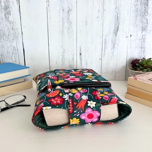 May include: A fabric book sleeve featuring a dark green background with a colorful floral pattern of pink, red, yellow, and white flowers. The sleeve has a snap button closure on a front flap and contains a thick book with visible pages.