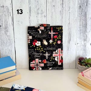 May include: A rectangular fabric sleeve featuring a black background with a repeating pattern of red and white flowers, butterflies, and ornate crosses. The text Amazing Grace is printed in white script throughout the design, which includes a snap closure.