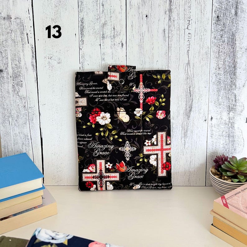 May include: A rectangular fabric sleeve featuring a black background with a repeating pattern of red and white flowers, butterflies, and ornate crosses. The text Amazing Grace is printed in white script throughout the design, which includes a snap closure.