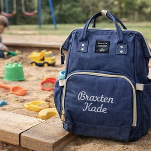 May include: A navy blue backpack with a top handle and a front zippered pocket. The name Braxten Kade is embroidered in white script on the front pocket. A black rectangular label with the text LEQUEEN and a crown icon is centered at the top.