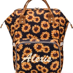 May include: Black backpack with a sunflower pattern. The bag has a brown handle and trim, and the name "Alexis" is printed on the front in a cursive font. The bag has multiple pockets and zippers.