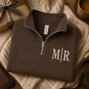 May include: A dark brown quarter-zip pullover featuring a silver metal zipper and a white embroidered monogram on the chest with the letters M and R separated by a vertical line. The garment has a high collar and a white interior neck label.