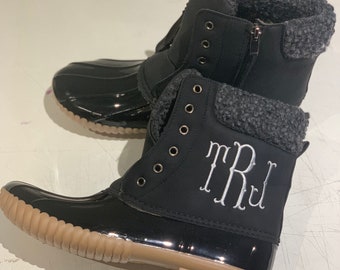 Op de afbeelding: Zwarte winterlaarzen met een bruine rubberen zool en een grijze fleecevoering. De laarzen hebben een rits aan de zijkant en een wit monogram met de tekst "TRJ".