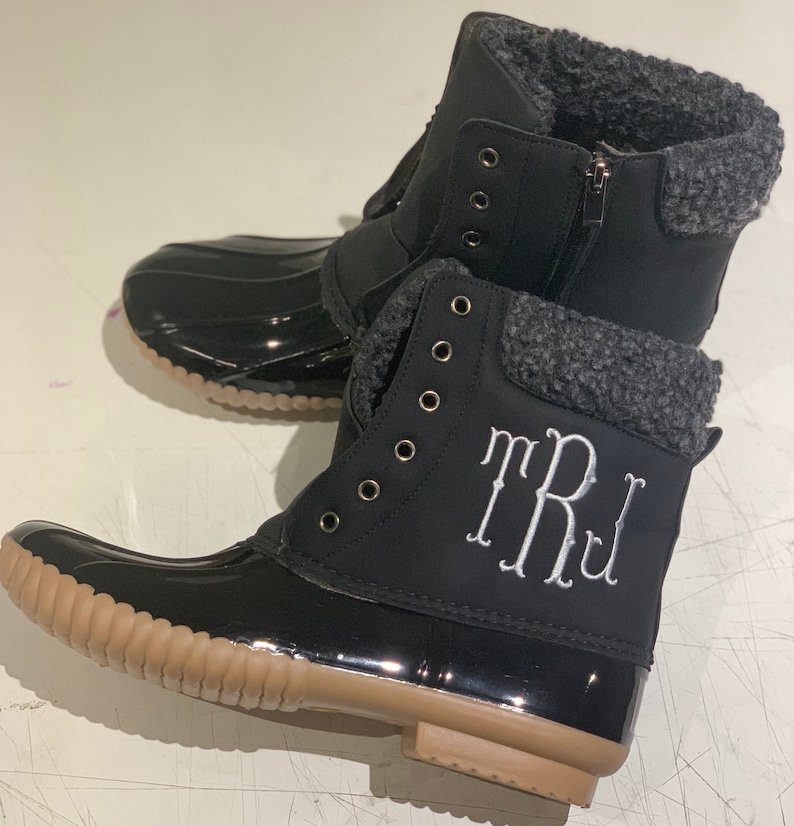 Op de afbeelding: Zwarte winterlaarzen met een bruine rubberen zool en een grijze fleecevoering. De laarzen hebben een rits aan de zijkant en een wit monogram met de tekst "TRJ".