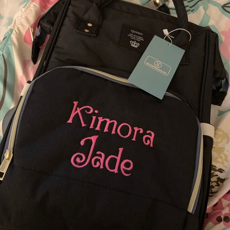 K&ouml;nnte beinhalten: Schwarze Wickeltasche mit goldenen Rei&szlig;verschl&uuml;ssen und grauem Besatz. Die Tasche ist mit einer personalisierten Stickerei versehen, die "Kimora Jade" lautet.