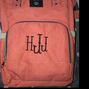 Puede incluir: Una bolsa de pa&ntilde;ales de color coral con un bolsillo frontal con cremallera y un monograma bordado en negro "HJL".