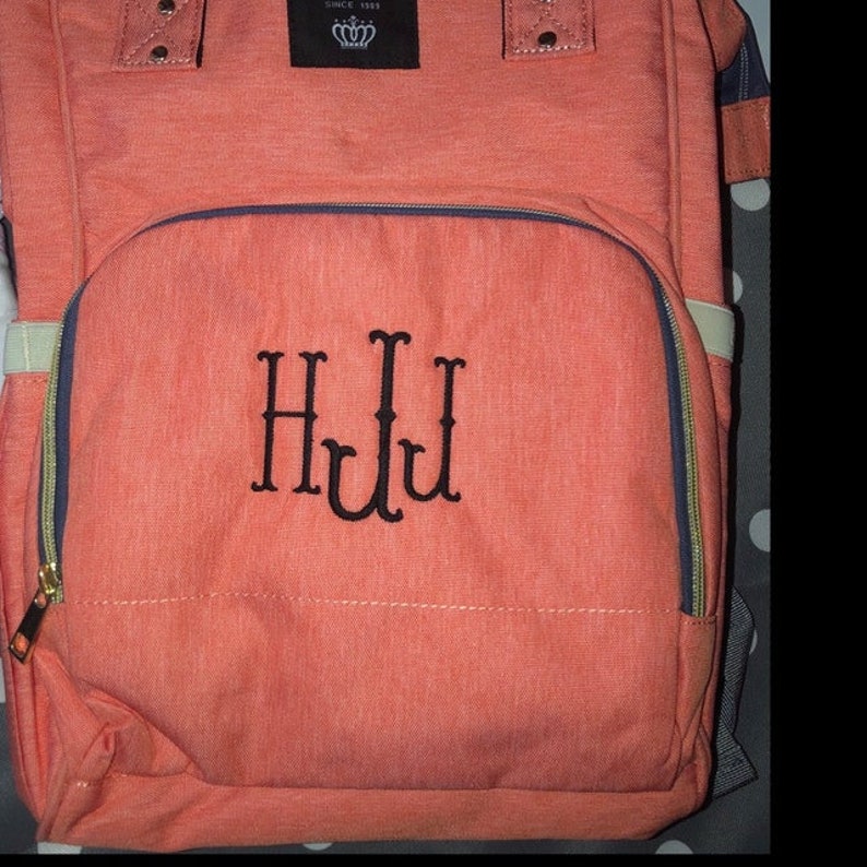 Puede incluir: Una bolsa de pa&ntilde;ales de color coral con un bolsillo frontal con cremallera y un monograma bordado en negro "HJL".