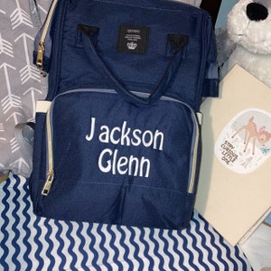 May include: A navy blue diaper bag with a white zipper and a white embroidered name "Jackson Glenn". The bag has a top handle and adjustable straps. The bag is sitting on a white and blue striped surface.