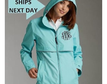 May include: A light blue hooded rain jacket with a zipper closure and front pockets. The jacket features a black monogram on the chest and a reflective stripe. The text "SHIPS NEXT DAY" is in a light blue arrow.