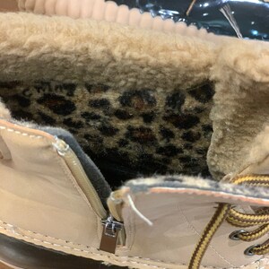 Op de afbeelding: Close-up van de binnenkant van een beige winterlaars met een zwarte rubberen zool. De laars heeft een rits aan de zijkant en een bruin en gele vetersluiting. De binnenkant van de laars is gevoerd met een zachte, bruine imitatiebont en heeft een binnenzool met luipaardprint.