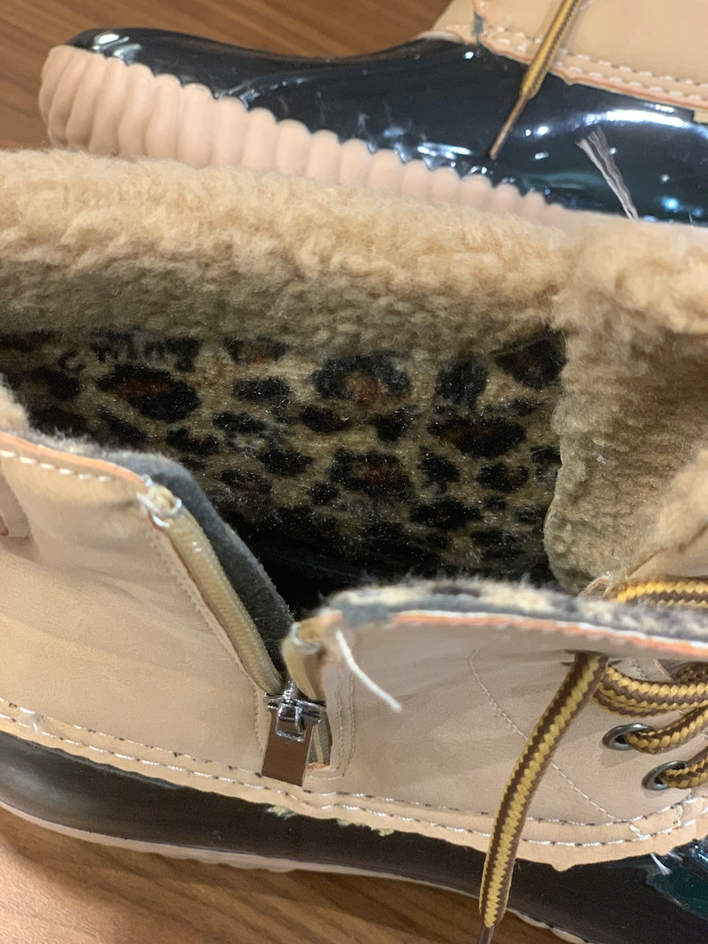 Op de afbeelding: Close-up van de binnenkant van een beige winterlaars met een zwarte rubberen zool. De laars heeft een rits aan de zijkant en een bruin en gele vetersluiting. De binnenkant van de laars is gevoerd met een zachte, bruine imitatiebont en heeft een binnenzool met luipaardprint.