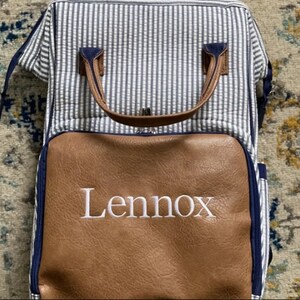 May include: A blue and white striped diaper bag with a brown leather bottom. The bag has a zipper closure and a handle. The bag is personalized with the name "Lennox" in white embroidery.