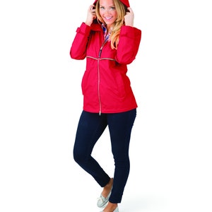 May include: A red hooded jacket with a zipper closure. The jacket has a white zipper pull and a white stripe down the front. The model is wearing blue jeans and white shoes.