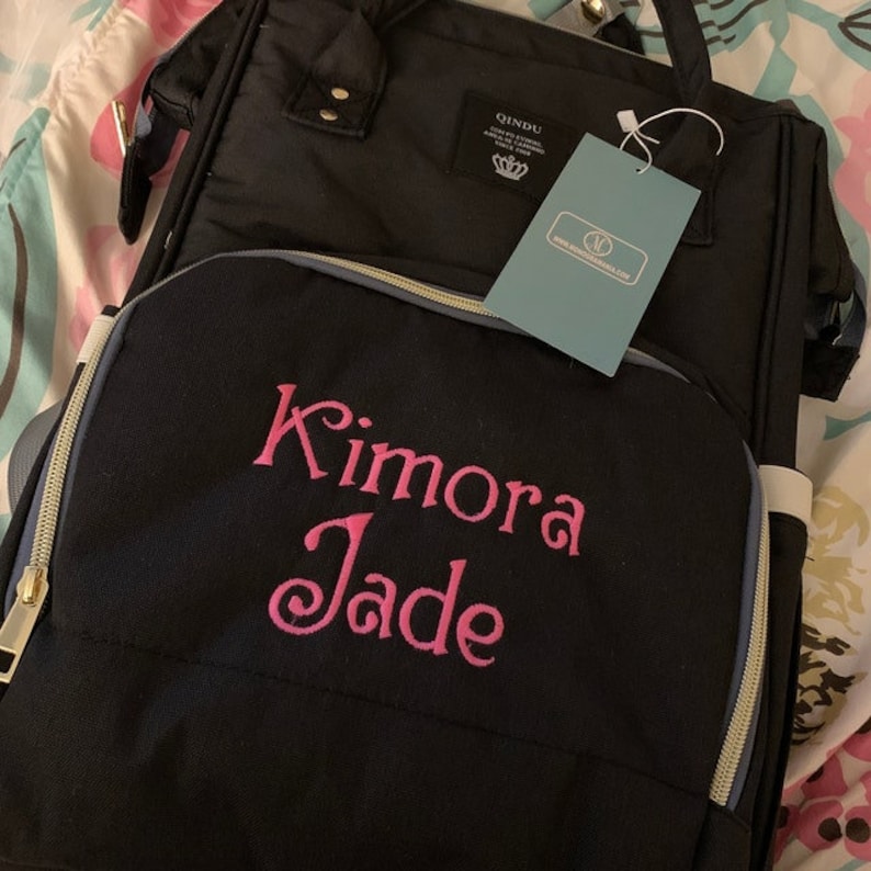 Puede incluir: Bolso de pa&ntilde;ales negro con cremalleras doradas y ribete gris. El bolso tiene un bordado personalizado que dice "Kimora Jade".
