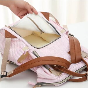 May include: A pink and white striped diaper bag with brown leather straps and a zipper closure. The bag has a padded interior and multiple pockets.