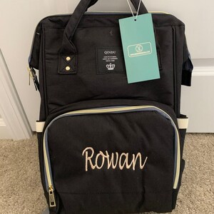 May include: Black diaper bag backpack with gold accents and a zippered pocket. The bag has a white strap and the name "Rowan" embroidered on the front in gold thread.