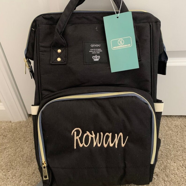 May include: Black diaper bag backpack with gold accents and a zippered pocket. The bag has a white strap and the name "Rowan" embroidered on the front in gold thread.