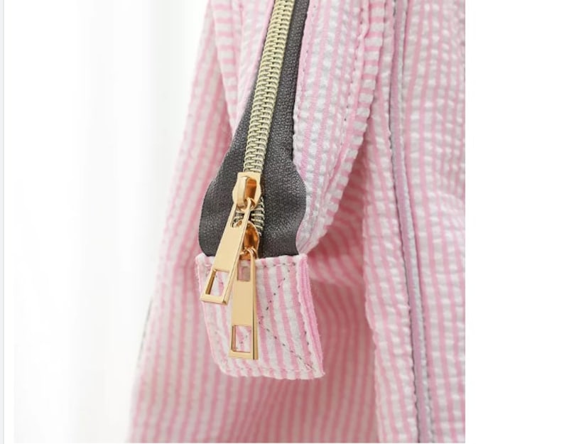 May include: Close-up of a pink and white striped zipper pull on a bag. The zipper pull is gold metal with a gray zipper tape.